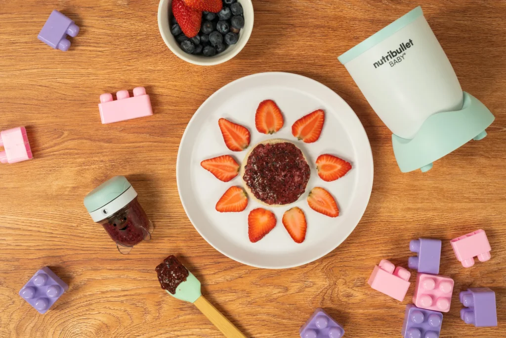Berry Chia Spread, una nutribullet® Baby junto con un contenedor de guardado, un plato con pan y el spread encima decorado para bebés, y juguetes de bebé en la mesa