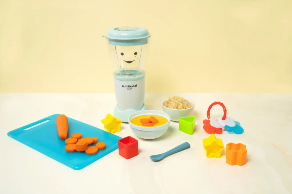 Sopa de Pollo, Arroz y Zanahoria, una nutribullet® Baby junto con ingredientes picados y juguetes para bebé. En el centro, un bowl de la sopa.