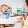 Bebé en su silla para comer siendo alimentado por su mamá, con la nutribullet® Baby Steam & Blend en la encimera de la cocina en el fondo