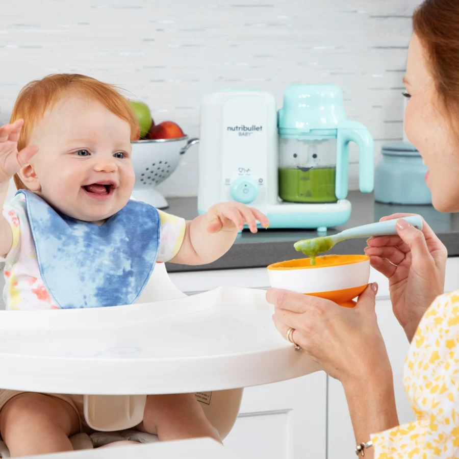 Bebé en su silla para comer siendo alimentado por su mamá, con la nutribullet® Baby Steam & Blend en la encimera de la cocina en el fondo