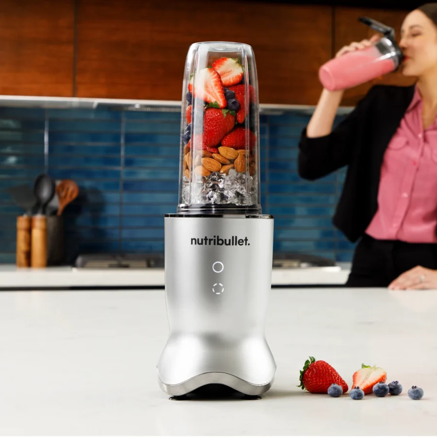 nutribullet® Ultra en color silver con ingredientes en el vaso en una cocina, mujer en el fondo tomando un smoothie con un vaso con tapa To-Go