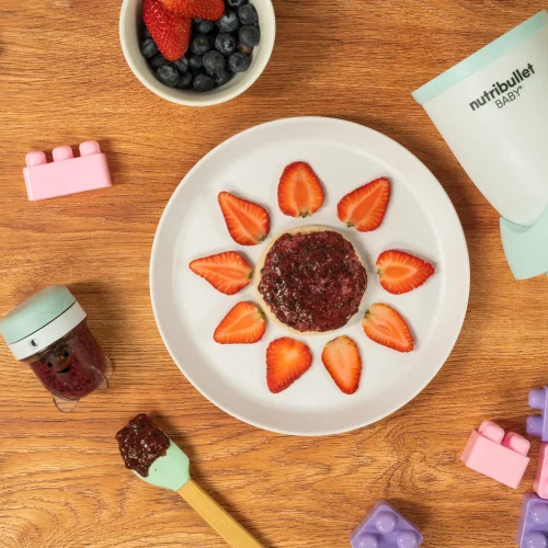 Berry Chia Spread, una nutribullet® Baby junto con un contenedor de guardado, un plato con pan y el spread encima decorado para bebés, y juguetes de bebé en la mesa