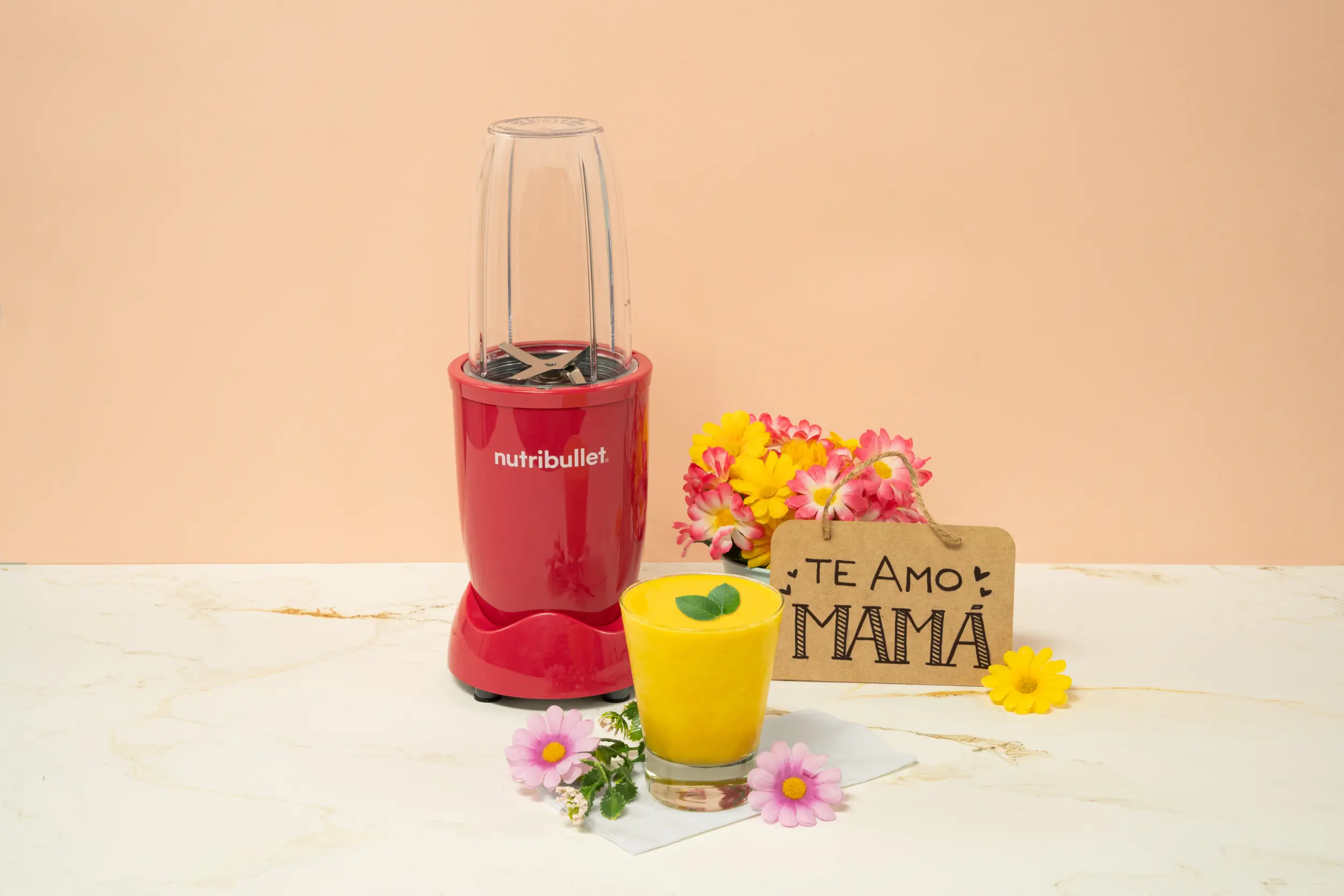 Smoothie Sunshine para mamá, una nutribullet® 600W Rubí de la Colección Glossy junto con flores, un letrero que dice ‘Te amo, Mamá’ y un vaso del smoothie