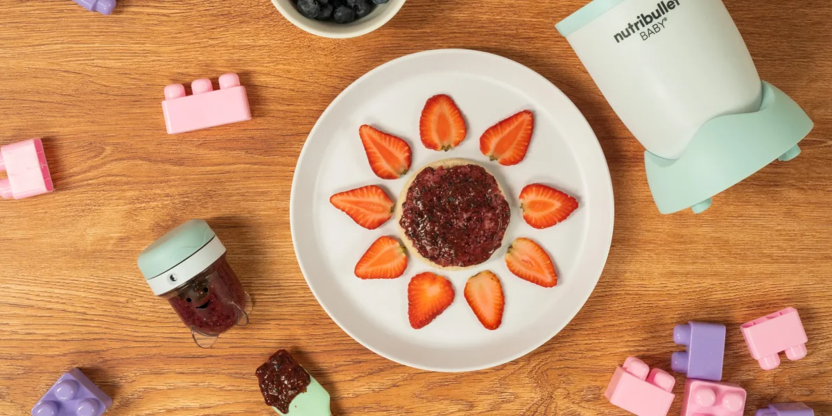 Berry Chia Spread, una nutribullet® Baby junto con un contenedor de guardado, un plato con pan y el spread encima decorado para bebés, y juguetes de bebé en la mesa